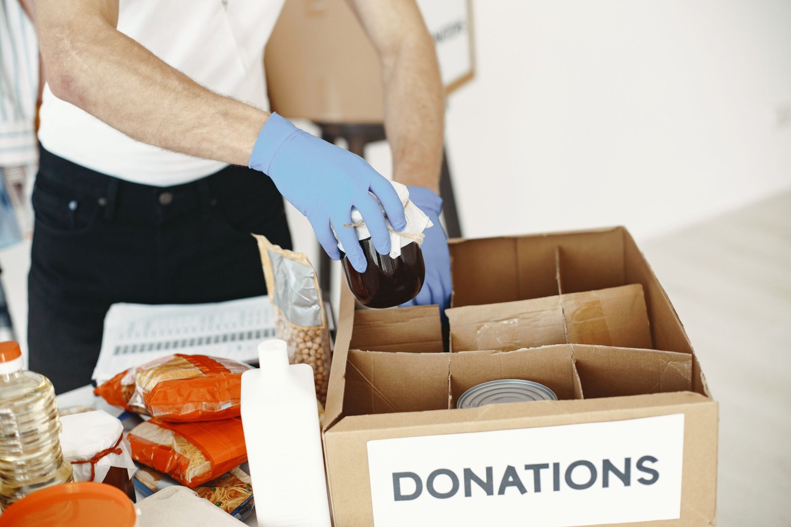 A volunteer with gloves packs canned goods and supplies in a donation box.